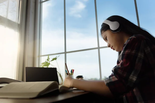 Female student wears headphones sitting at table to learning and note, using laptop for homework during covid19 pandemic and lock down, Social Distance, New life of Student on internet. Mockup screen.