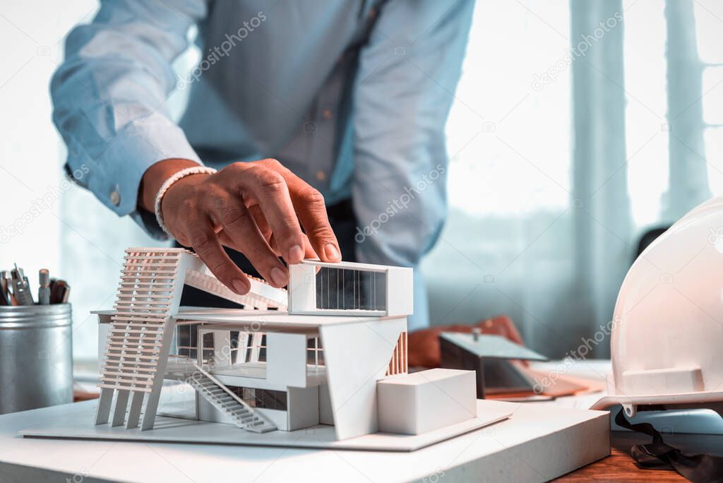 Hand of Asian architect-designer holding the part of the model of modern box house while thinking about concept of building and construction project in the future. Focusing on his hand holding model.
