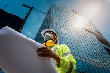 İnşaat mühendisleri, inşaat alanında maske ve güvenlik kaskı takmış ellerinde planlar tutuyorlardı. İnşaat müfettişi. İnşaat sahasının çizimi ve inşaatın iş akışı