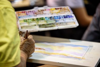 Geniş stüdyodaki diğer öğrencilerle birlikte resimlerine suluboya resim yapan yaşlı kadının eli. Yaşlılar için sanat aktiviteleri.