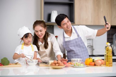 Asyalı bir aile kaseye yumurta atıyor ve kız beyaz masada yumurta çırpıyor ve anne ve babasıyla evde yemek pişirirken baba da akıllı telefonuyla selfie çekiyor..