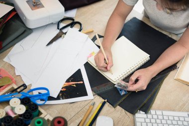 Masada kumaş ve terzinin kesim ve tasarım stüdyosunda ekipman olan bir notla birlikte kadın terzinin elinin üst görüntüsü. Notada çok sığ odak noktası