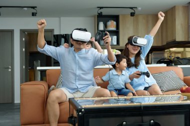 Asyalı bir aile evdeki oturma odasında sanal gerçeklik VR kulaklığı kullanarak oyun oynuyor, neşeli bir aile birlikte eğleniyor. Aile zamanı kavramı