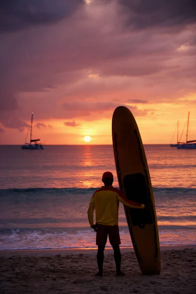 夕日の景色を眺めながらビーチでサーフと一緒に立っているビーチライフガードのシルエットと背景にヨット — ストック写真