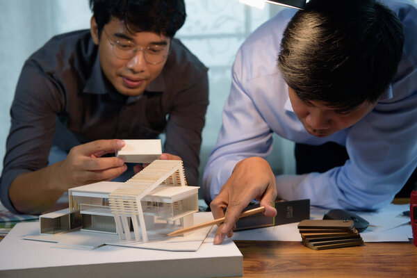Asian architects discussing with designer or civil engineer about concept of building design and construction project for modern box house. Focusing on the house model.