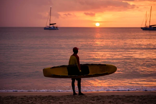 背景の夕日とヨットを見ながら 空のビーチにサーフボードで一人で立っているビーチライフガードのシルエット コロナウイルス 観光地や観光損失への経済的影響 — ストック写真