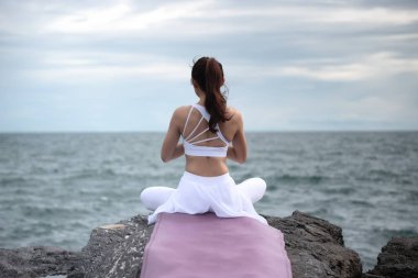 Sukhasana egzersizinde yoga yapan Asyalı genç bir kadın denize bakarken....