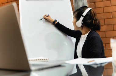 Asyalı öğretmen online masada oturuyor ya da sınıftaki öğrencilere beyaz tahtayla ders veriyor. Modern eğitim, karantina eğitimi, Çevrimiçi eğitim kavramı.