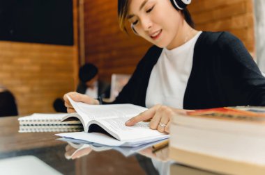 Asyalı burslu öğrenci ders notlarını dinlemek için kulaklık takarken masa başında kitap okuyor. Eğitim araştırması ve kendini geliştirme. Eğitimsel fırsat kavramı.