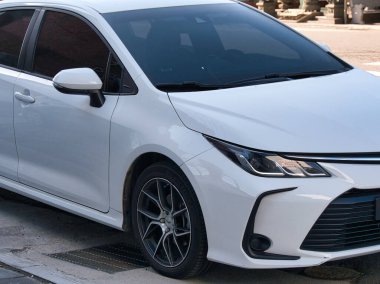 Şehir merkezinde asfalt bir sokağa park edilmiş modern bir beyaz sedan, gösterişli tasarım ayrıntıları ve koyu renkli pencerelerle, çağdaş bir tarz ve günlük işe gidiş-dönüş hissini aktarıyor..