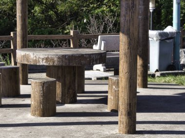 Açık havada beton masa ve tabureler doğal bir ortamda, taklit ahşap dokuya sahip ve yemyeşil yapraklarla çevrili, kırsal ve huzurlu bir atmosferi taşır..