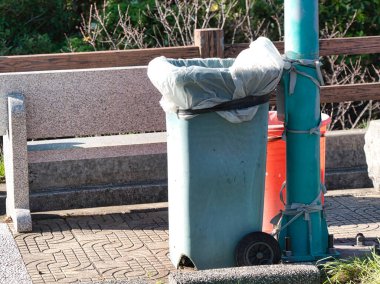 Beyaz çizgili bir deniz mavisi çöp tenekesi kaldırımlı bir alanda granit bir bankın yanında duruyor, arka planda ahşap bir çit ve yeşil yapraklar, sıradan ama gerekli bir şehir manzarasını tasvir ediyor..