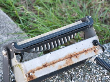 Paslanmış ve yıpranmış bir endüstriyel ışık armatürünün yakından görüntüsü, arka planda yeşil çimenler olan bir çakıl ve beton yüzey üzerinde oturan, yaşlılık ve ihmalkârlık duygusunu aktaran.