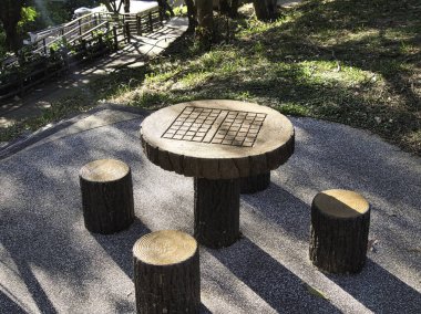 Açık hava beton masa ve ağaç gövdelerine benzemesi için tasarlanmış sandalyeler, güneşli bir park ortamında masa oyunları için dama desenleri, dinlenme ve dinlenmeye davetiye çıkarıyor..