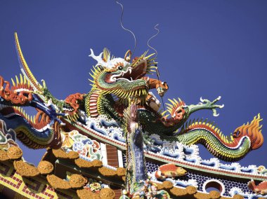 Canlı, renkli bir ejderha heykeli açık mavi gökyüzünün altında bir tapınak çatısını süslüyor. Geleneksel mimarinin gücünü ve iyi talihini simgeliyor..