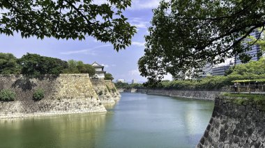 Japonya 'nın Osaka Kalesi' ni çevreleyen hendeğin manzarası. Çekimi çevreleyen yemyeşil ağaçlar ve sakin suya yansıyan tarihi taş duvarlar..