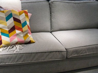 Tercümesi Sanela. Renkli geometrik desenli, gri renkli bir kanepe fırlatma yastığı ve beyaz püsküllü bir battaniye..