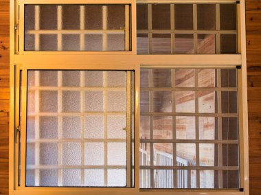 Metal güvenlik ızgarası ve ahşap bir duvara yapıştırılmış buzlu cam paneller olan bir mesken penceresine yakın plan. Ilık iç aydınlatma desenli cam ve çerçeveyi aydınlatır.