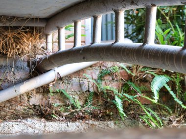 Köprü ya da yürüyüş yolunun altındaki metal destek yapısının yakın görüntüsünde yıpranmış boya, PVC boru ve doğal ışık altında eskimiş tuğla bir duvara karşı büyüyen yeşil eğrelti otları yer alıyor..