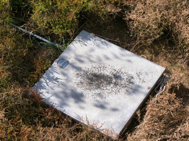 Yıpranmış gümüş kapak, doğal bir çevrede endüstriyel bozulma hissi uyandıran koyu bir radyal leke taşıyan metal yüzeyi vahşi kıyı çalılıklarına gömülü..