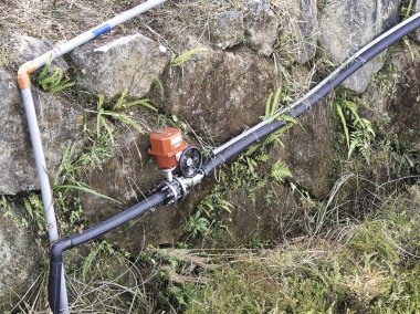 Bir motorlu kırmızı aktüatör kapakçığı, kırsal bir doğal ortamda modern kullanım altyapısını vurgulayan engebeli bir taş setine karşı açık hava boru hattı sistemine entegre bir şekilde duruyor..