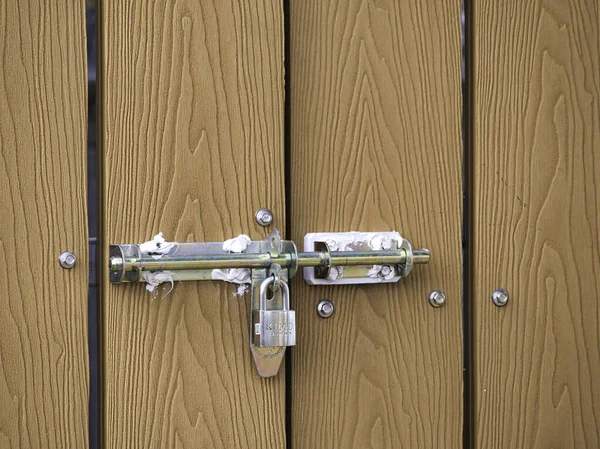 Gümüş asma kilit ile metal sürgülü kilit, açık havada kahverengi sahte ahşap bir çiti güvence altına alır. Güneşli bir günde güçlü bir güvenlik, koruma ve konut gizliliği hissi verir..