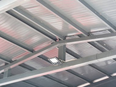 Modern bir LED projektör ışığı bükülmüş metal bir çatının altındaki gri çelik bir kirişe monte edilir ve güvenlik ve işlevsel tasarım odaklı bir endüstriyel tesis için gerekli aydınlatmayı sağlar..