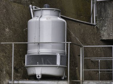 Yıpranmış beton bir duvara karşı yüksek bir platforma monte edilmiş büyük gri endüstriyel soğutma kulesi kamu hizmetleri altyapısı ve mekanik verimliliği gösteriyor..