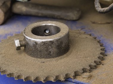 Tozlu bir endüstriyel metal dişli çıkıntısı ahşap talaşı ve kir ile kaplı mavi bir tezgahta duruyor. Bu da atölye ortamında ağır makinelerin çalışmasını sembolize ediyor..