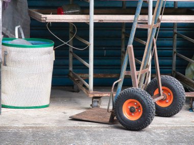 Parlak turuncu tekerlekleri olan yıpranmış mavi bir el kamyonu kırsal bir sokakta beyaz bir sepetin yanında duruyor..