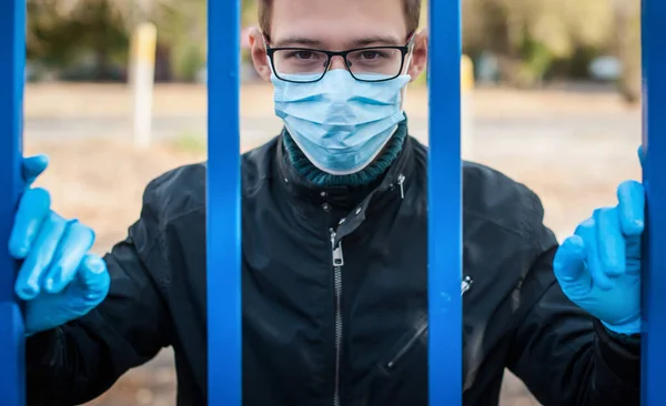 A man in a medical mask and gloves behind a metal bars in the street. Quarantine and isolation concept. Coronavirus and covid-19 flu virus