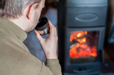 Evdeki şöminenin önünde termostan sıcak çay içen bir adam. Yakın plan. Kır evi, kış, dinlenme, rahatlama. Konu odak dışı. 