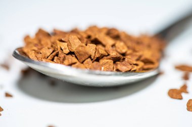 Temiz beyaz bir zemin üzerine yerleştirilmiş koyu kahverengi kahve taneleriyle dolu bir kaşığın detaylı makro fotoğrafı, doku ve aromayı ön plana çıkarıyor..