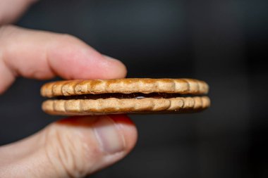 Kakaolu bisküvi sandviçi tutan bir elin detaylı makro fotoğrafı zengin çikolata dolgusunu, parçalanmış dokusunu ve iştah açıcı görünümünü gözler önüne seriyor. Yemek reklamı, ürün ambalajı ve tatlı promosyonları için ideal..