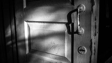 Eski ahşap kapının yarı açık, dramatik ışık ve gölgelerle aydınlatılmış tek renkli bir fotoğrafı. Klasik doku, tutuş ve gizemli atmosfer gerilim ve tarih duygusu ile sinematik, karamsar bir kompozisyon yaratır..