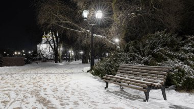 Karla kaplı yolu olan sakin bir gece parkı sahnesi, eski tarz banklar ve parlayan fenerler. Sakin atmosferi olan kış şehirleri.