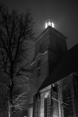 Almanya, Berlin, 19 Aralık 2025. Dar, eski kasaba sokakları arasında yükselen tarihi bir kulenin gece manzarası. Sıcak yapay ışık derin gölgeler ve karanlık cephelerle çelişiyor dramatik ve sinematik bir atmosfer yaratıyor..