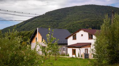 Yeşil bitki örtüsü ve ağaçlarla çevrili kırmızı çatılı küçük bir kırsal ev, bir dağ manzarasına karşı kurulmuş. Sahne huzurlu kırsal yaşamı ve doğayla uyumu gösteriyor. Kırsal yaşam tarzı, seyahat ve geleneksel mimari kavramı.