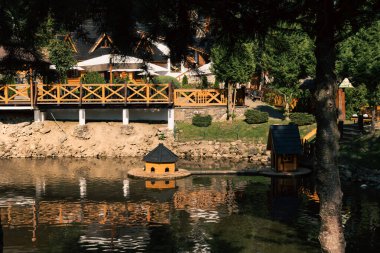 Güneşli bir günde ağaçlarla ve orman tepeleriyle çevrili, şemsiyeli ahşap bir restoran terası. Suda yansımaları ve sıcak doğal ışığı olan bir dağlık bir tatil beldesinde rahat bir açık hava yemek alanı..