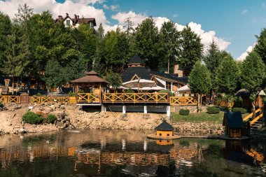 Güneşli bir günde ağaçlarla ve orman tepeleriyle çevrili, şemsiyeli ahşap bir restoran terası. Suda yansımaları ve sıcak doğal ışığı olan bir dağlık bir tatil beldesinde rahat bir açık hava yemek alanı..