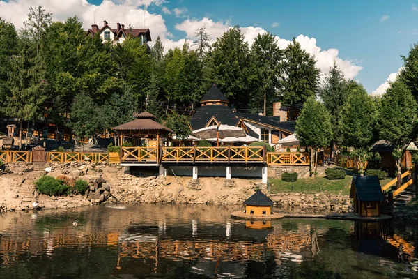 Güneşli bir günde ağaçlarla ve orman tepeleriyle çevrili, şemsiyeli ahşap bir restoran terası. Suda yansımaları ve sıcak doğal ışığı olan bir dağlık bir tatil beldesinde rahat bir açık hava yemek alanı..