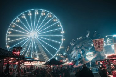 Parlak dönme dolap, şenlikli ışıklar ve cazibelerle dolu canlı bir panayırın üzerinde gece aydınlandı. Kalabalık insanlar ve renkli tezgahlar enerjik ve neşeli bir atmosfer yaratıyor..