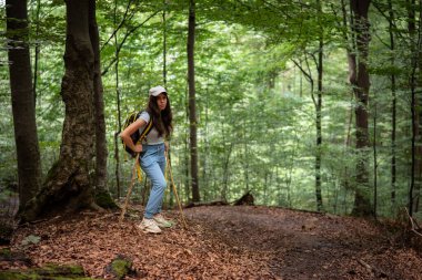 Yeşillik bir ormanda yürüyüş yapan genç bir kadın, doğal bir patika boyunca yürürken tahta bir baston kullanıyor. Aktif yaşam tarzı konsepti, açık hava macerası, eko turizm ve doğa ile bağlantı.