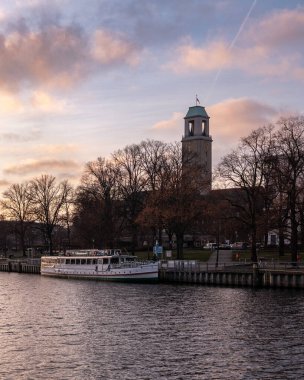 Gün batımında şehrin içinden akan sakin bir nehrin geniş panoramik görüntüsü, tarihi binalar, set boyunca uzanan ağaçlar ve suya yansıyan yumuşak yansımalar. Sıcak akşam ışığı ve bulutlar barışçıl ve atmosferik şehir manzarası yaratır..