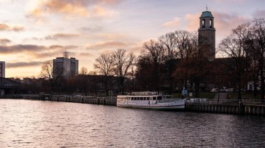 Gün batımında şehrin içinden akan sakin bir nehrin geniş panoramik görüntüsü, tarihi binalar, set boyunca uzanan ağaçlar ve suya yansıyan yumuşak yansımalar. Sıcak akşam ışığı ve bulutlar barışçıl ve atmosferik şehir manzarası yaratır..