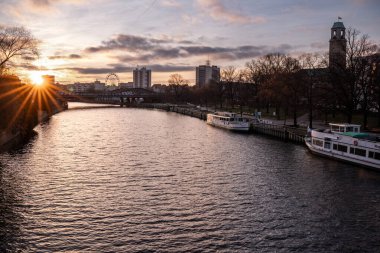Gün batımında şehrin içinden akan sakin bir nehrin geniş panoramik görüntüsü, tarihi binalar, set boyunca uzanan ağaçlar ve suya yansıyan yumuşak yansımalar. Sıcak akşam ışığı ve bulutlar barışçıl ve atmosferik şehir manzarası yaratır..