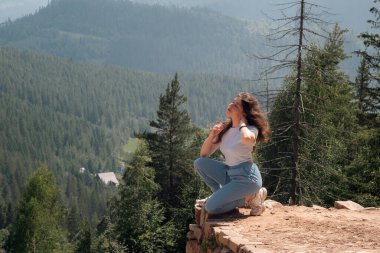 Diz çöküp orman tepeleriyle çevrili temiz dağ havasının tadını çıkaran genç bir kadın. Özgürlükle, rahatlıkla ve doğayla bağlantıyla yazlık açık hava yaşam tarzı konsepti. Manzaralı kırsal alan ve huzurlu seyahat atmosferi.