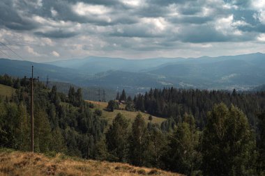 Yoğun yeşil ormanları, yuvarlanan tepeleri ve dramatik bulutlu bir gökyüzünün altındaki uzak köyün geniş panoramik manzarası. Seyahat, turizm, çevre, sürdürülebilirlik ve açık hava arka plan kavramları için uygun doğal yaz manzarası.
