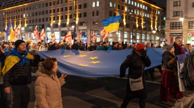 Berlin, Almanya - 24 Şubat 2026: Göstericiler Ukrayna ve Ukrayna halkını destekleyen bir miting sırasında gece aydınlık bir şehir kavşağında yürüyorlar. Büyük bir Avrupa Birliği bayrağı kış örtüsü katılımcıları tarafından yatay olarak taşınır