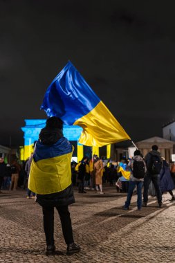 BERLİN, Almanya FEBRUARY 24, 2026: Ukrayna bayrağına sarılmış bir gösterici, Ukrayna ve Ukrayna halkına destek için Rusya 'nın tam ölçekli işgalinin yıldönümünde düzenlenen mitingde Brandenburg Kapısı yakınlarında duruyor. Büyük bir mavi-yel